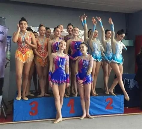 Young female gymnasts in sparkly leotards standing on a podium with gold medals, celebrating competition results indoors